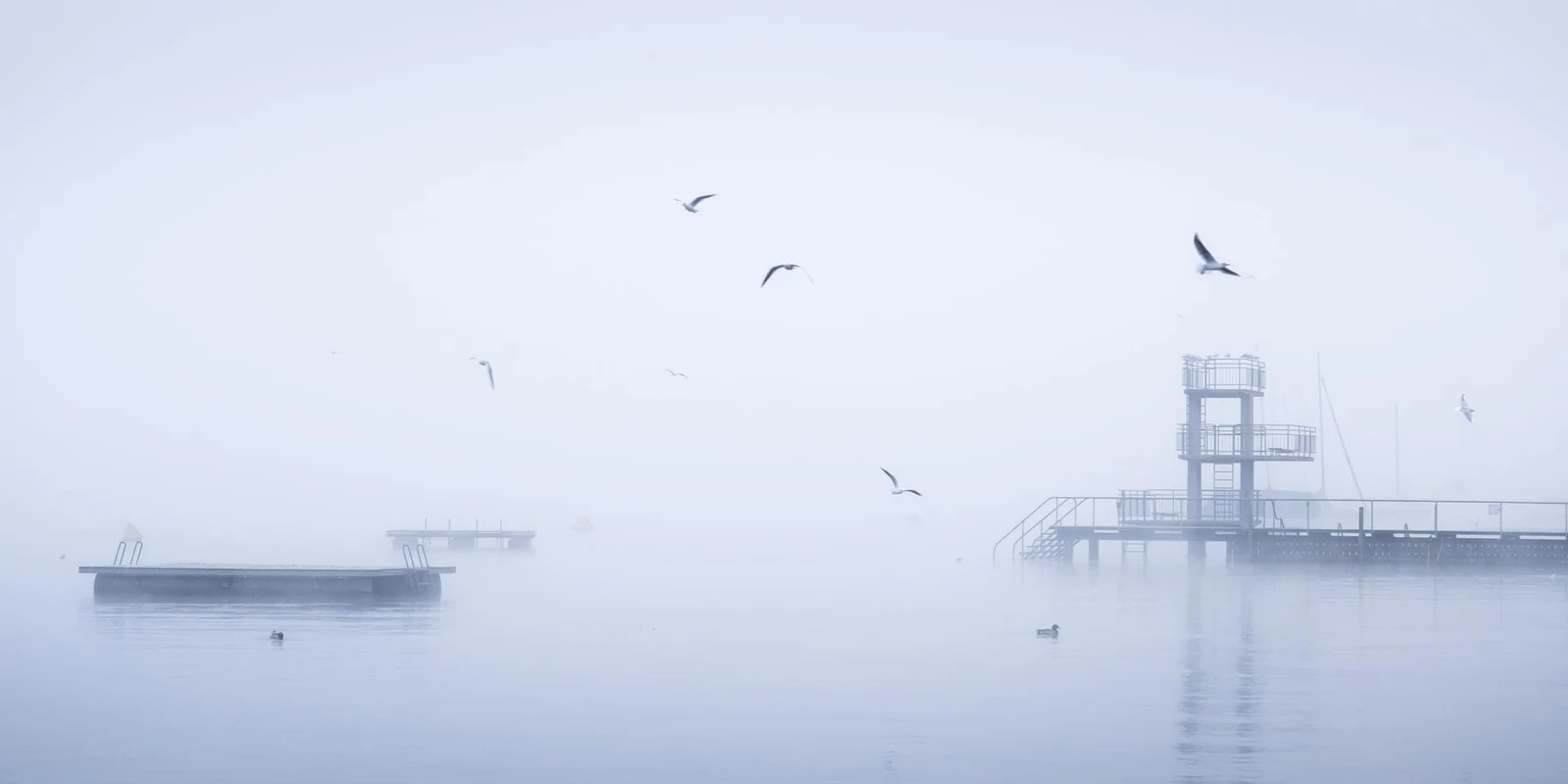 Fine art print of a minimalist lake scene in heavy mist. This high-key photograph captures a geometric pier and a complex diving platform dissolving into the white fog.